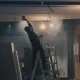 Remont wnętrza: pracownik na drabinie montuje sufit, w tle materiały budowlane i lampy.