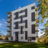 Nowoczesny budynek mieszkalny z balkonami i geometrycznym wzorem na elewacji, otoczony zielenią.
