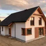 Nowoczesny dom z białym tynkiem, drewnianymi elementami i czarnym dachem na tle zachodzącego słońca.