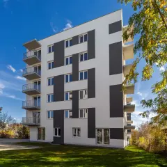 Nowoczesny budynek mieszkalny z balkonami i geometrycznym wzorem na elewacji, otoczony zielenią.