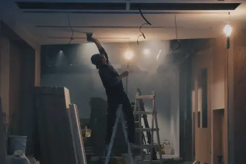 Remont wnętrza: pracownik na drabinie montuje sufit, w tle materiały budowlane i lampy.