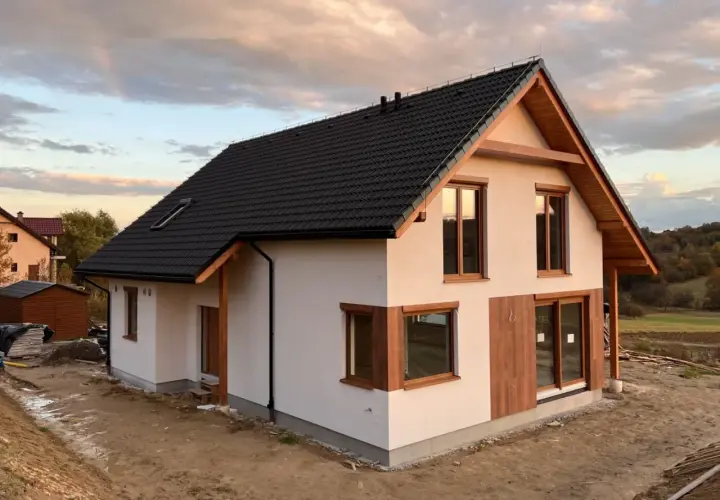Nowoczesny dom z białym tynkiem, drewnianymi elementami i czarnym dachem na tle zachodzącego słońca.